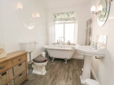 A bathroom with a clawfoot bathtub a toilet a pedestal sink a wooden cabinet with baskets and a window with a floral blind at Pottergill in Windermere