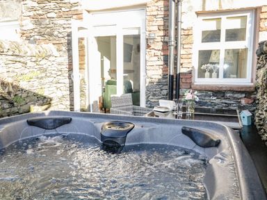 An outdoor hot tub with bubbling water next to a glass table with flowers and glasses against a stone wall at Pottergill in Windermere