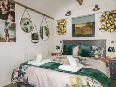 A bedroom with a bed with floral bedding green pillows and towels on top with mirrors flower decorations and paintings on the wall at High Rigg Shepherd's Hideaway in Brampton Cumbria