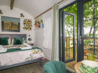 A bedroom with a bed floral bedding robes on hooks and glass doors opening to a deck with chairs at High Rigg Shepherd's Hideaway in Brampton Cumbria