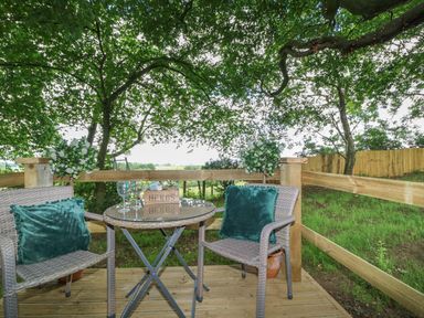 An outdoor seating area with two chairs and a round glass table on a wooden deck surrounded by trees and a wooden fence at High Rigg Shepherd's Hideaway in Brampton Cumbria