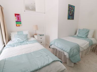 A bedroom with two single beds with light blue bedding a bedside table with a lamp and books and abstract paintings on the walls at 1A Chantry Place in Morpeth