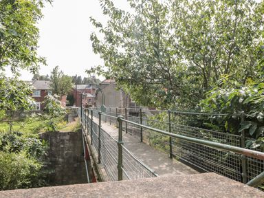 A narrow pedestrian bridge with metal railings surrounded by trees and buildings at 1A Chantry Place in Morpeth