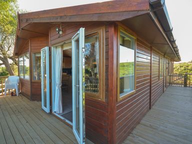 An exterior view of a wooden lodge with open glass doors leading to a deck with stacked plastic chairs at Ffrwd Lodge in Rhosneigr