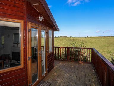 A wooden deck with railing outside a cabin overlooking grassy fields at Ffrwd Lodge in Rhosneigr