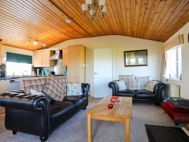 A living room with two black leather sofas and a wooden coffee table next to a kitchen area at Ffrwd Lodge in Rhosneigr