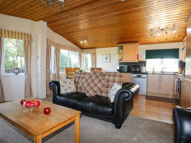 A living room with a black leather sofa and wooden coffee table next to a kitchen with wooden cabinets at Ffrwd Lodge in Rhosneigr