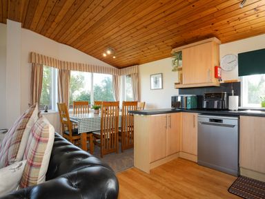A kitchen with wooden cabinets dining table with chairs and black leather sofa at Ffrwd Lodge in Rhosneigr