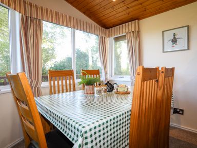 A dining area with a checkered tablecloth wooden chairs and large windows with striped curtains at Ffrwd Lodge in Rhosneigr