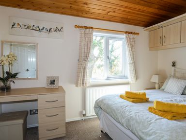 A bedroom with a bed towels on it a window with checked curtains and a desk with a mirror and flowers at Ffrwd Lodge in Rhosneigr