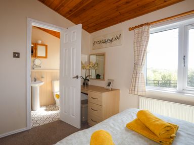 A bedroom with a bed and yellow towels a dresser and mirror a window with checked curtains and an open door to a bathroom at Ffrwd Lodge in Rhosneigr
