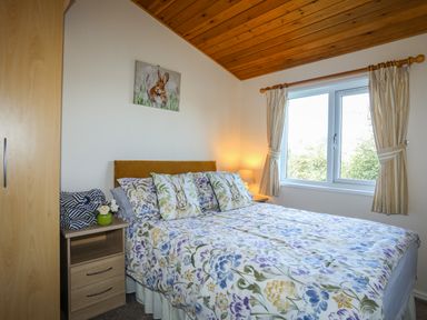 A bedroom with a double bed with floral bedspread pillows with rabbits bedside table lamp window with curtains and wooden wardrobe at Ffrwd Lodge in Rhosneigr