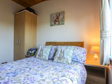 A bedroom with a bed with floral and rabbit pillows a wooden wardrobe a nightstand with a lamp and a rabbit painting above the bed at Ffrwd Lodge in Rhosneigr