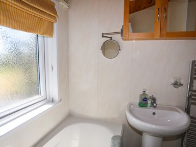 A bathroom with a bathtub a sink a wall-mounted mirror and a window with a yellow blind at Ffrwd Lodge in Rhosneigr