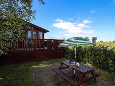 An outdoor wooden picnic table with a green and white striped umbrella next to a wooden cabin in a grassy area at Ffrwd Lodge in Rhosneigr