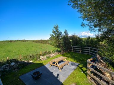 An outdoor seating area with a wooden picnic table on a paved patio surrounded by grass and fencing at Ffrwd Lodge in Rhosneigr