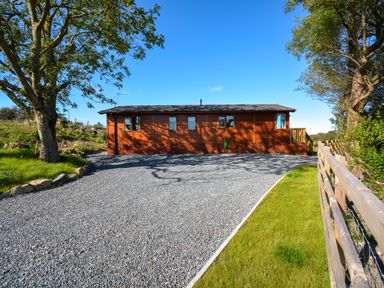 A wooden building with gravel driveway green grass and trees at Ffrwd Lodge in Rhosneigr
