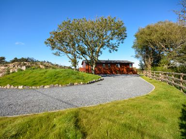 A gravel driveway leading to a wooden house with trees and a wooden fence at Ffrwd Lodge in Rhosneigr