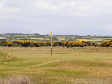 A golf course with a yellow flag and grassy hills with distant houses and fields in the background at Ffrwd Lodge in Rhosneigr