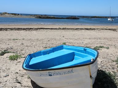 A small white and blue boat on a sandy beach with rocks and a sailboat in the water at Ffrwd Lodge in Rhosneigr