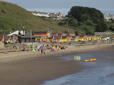 A beach with people, colorful beach huts, grassy hills, and houses in the background at Jonstone Pines in Cayton Bay near Scarborough