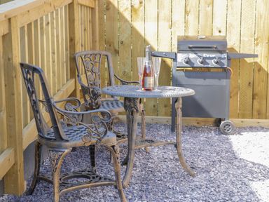 An outdoor seating area with two metal chairs and a round table with a bottle and two glasses next to a barbecue grill at Bluebell Wood in Brandesburton