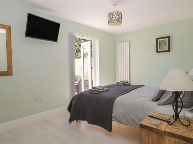 A bedroom with a bed covered in gray bedding a bedside table with a lamp and remote a wall-mounted television and open doors to an outdoor area at Bluebell Wood in Brandesburton