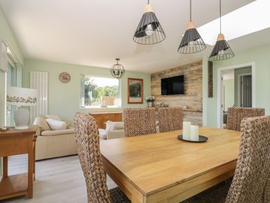 A dining area with a wooden table and wicker chairs adjacent to a living room with sofas a wall-mounted TV and large windows at Bluebell Wood in Brandesburton