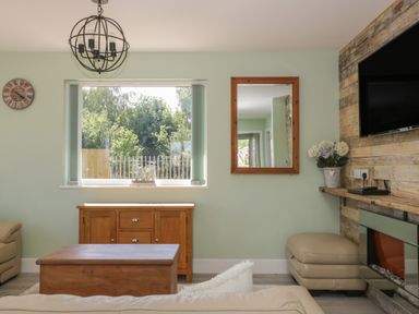 A living room with a wooden coffee table a window with vertical blinds a wall clock a mirror a wooden sideboard a wall-mounted television a flower pot and a fireplace at Bluebell Wood in Brandesburton