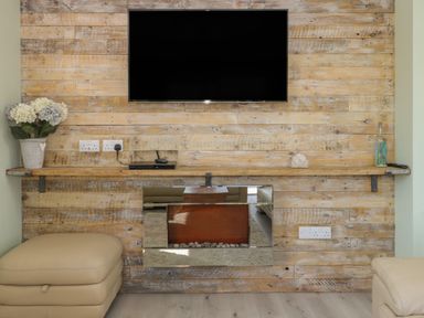 A wooden wall with a mounted television a wooden shelf with decorative items and a beige ottoman at Bluebell Wood in Brandesburton