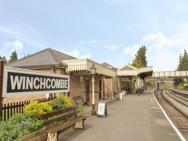A train station platform with benches a sign reading Winchcombe and a brick building with canopies at Thimble Cottage Winchcombe