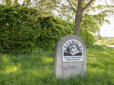 A stone sign with a sheep illustration and text cotswolds area of outstanding natural beauty beside a tree and hedge at Thimble Cottage in Winchcombe