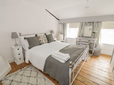 A bedroom with a bed with gray and floral pillows and a bedside table with a lamp at Thimble Cottage in Winchcombe