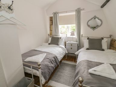A bedroom with two single beds a bedside table a lamp a window with curtains and a decorative mirror at Thimble Cottage in Winchcombe