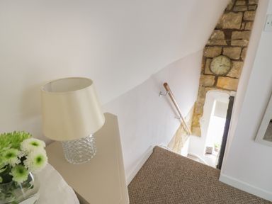 A staircase with a wooden handrail a wall clock exposed stonework and a lamp on a table at Thimble Cottage in Winchcombe