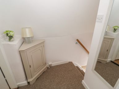 A staircase corner with a small cabinet a lamp a vase with flowers and a wall mirror at Thimble Cottage in Winchcombe