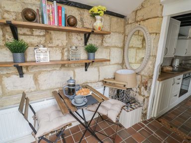 A small dining area with a wooden table set for coffee and croissants next to stone walls and shelves with books and plants at Thimble Cottage in Winchcombe