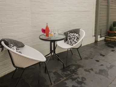 An outdoor patio area with two white chairs with cushions and throws around a round glass table with a bottle and glasses at Thimble Cottage in Winchcombe