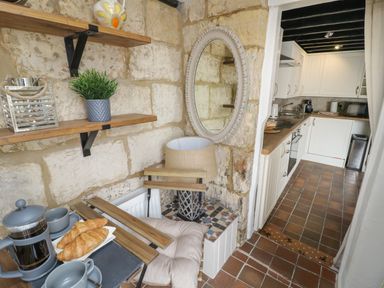 A small seating area with croissants on a tray and a lamp next to an open kitchen with white cabinets at Thimble Cottage in Winchcombe