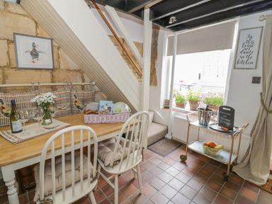 A dining area with a wooden table and white chairs under a staircase with cushions and a framed duck picture a trolley with wine and fruit by a window at Thimble Cottage in Winchcombe