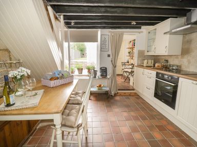 A kitchen with white cabinets a wooden table with chairs and a window showing outdoor plants at Thimble Cottage in Winchcombe