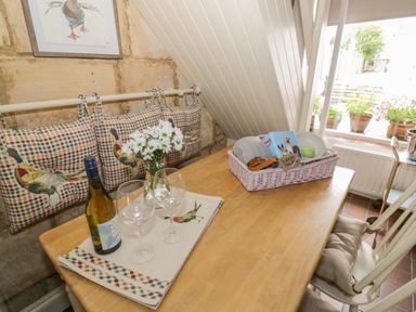 A wooden dining table with wine glasses flowers and a basket next to a window and chairs at Thimble Cottage in Winchcombe