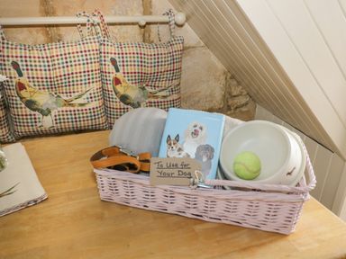 A basket with dog accessories on a wooden table in a room with cushioned bench at Thimble Cottage in Winchcombe
