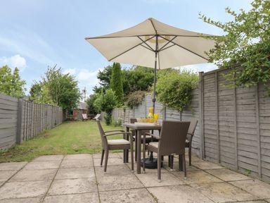 A patio area with a table and four chairs under a large umbrella in a garden with grass and fences on both sides at Thimble Cottage in Winchcombe