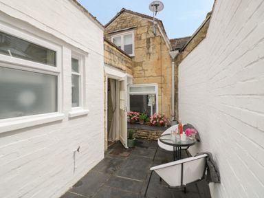 A small outdoor patio with black tile flooring white brick walls two chairs a round glass table and potted plants at Thimble Cottage in Winchcombe