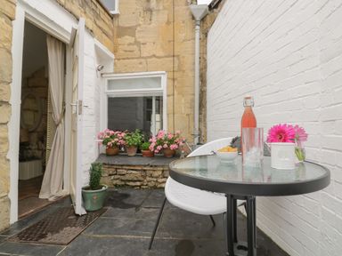 A small outdoor patio with a glass table set with a drink and flowers next to white walls and an open door with potted plants by the window at Thimble Cottage in Winchcombe