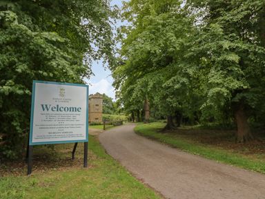 A driveway with a welcome sign for Sudeley Castle and Gardens surrounded by trees at Thimble Cottage in Winchcombe