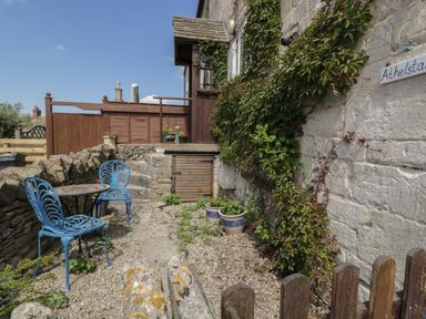 A small outdoor patio with two blue metal chairs a round table a stone wall and climbing plants on a cottage wall at Athelstan Cottage Chloe Mead near Stroud