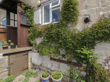 A stone wall with a window and climbing plants in pots and a wooden porch at Athelstan Cottage Chloe Mead near Stroud
