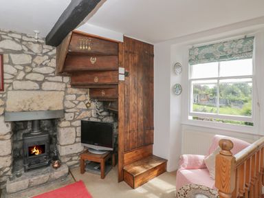 A stone wall with fireplace and wood stove a wooden staircase a small television a window with patterned blind and a cushioned chair at Athelstan Cottage in Chloe Mead near Stroud